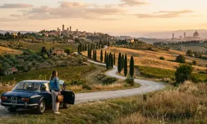 Donna con mappa accanto a un'auto d'epoca in un paesaggio collinare della Toscana da vedere, con cipressi e borghi all'orizzonte.