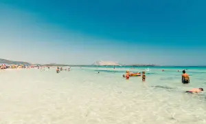 Spiaggia affollata in Sardegna, tra le zone più belle, con gente che nuota e si rilassa nell'acqua cristallina.
