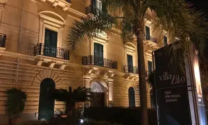 Villa Zito a Palermo, un'elegante dimora illuminata di notte, con palme che incorniciano la facciata e un cartello che annuncia "Dipinti delle collezioni permanenti".