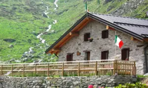 Rifugio in pietra con bandiera italiana, circondato da prati verdi e un ruscello. Un luogo ideale per rifugi val camonica facili.