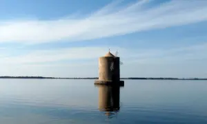Mulino solitario nella laguna di Orbetello, riflesso sull'acqua calma sotto un cielo azzurro con nuvole sottili.