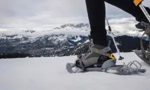 Una persona con ciaspole cammina sulla neve, con montagne innevate sullo sfondo.