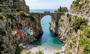 Un ponte ad arco domina un fiordo con case colorate aggrappate alla roccia. Il mare turchese invita a un bagno, mentre la strada panoramica, teatro del furore costiera amalfitana, accoglie bus e moto.