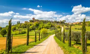 Strada sterrata fiancheggiata da cipressi in una gita in Toscana, con un borgo in collina sotto un cielo azzurro e nuvoloso.
