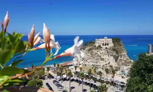 Fiori bianchi in primo piano, sullo sfondo il Santuario di Santa Maria dell'Isola a Tropea, un must per chi cerca cosa vedere in Calabria. Spiaggia affollata e mare azzurro.