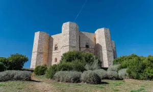 Castel del Monte, un'imponente fortezza ottagonale in pietra chiara, si erge maestoso sotto un cielo azzurro intenso, circondato da vegetazione mediterranea.