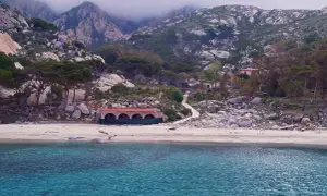 Spiaggia selvaggia con rocce e vegetazione, un edificio storico e montagne nebbiose sullo sfondo. Qui si trova l'isola di Montecristo.
