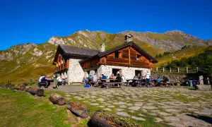 Un rifugio di montagna in pietra e legno, con persone sedute ai tavoli all'aperto, circondato da prati verdi e montagne imponenti sotto un cielo azzurro.