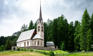 Chiesa di San Giovanni di Fassa, con campanile a cuspide, circondata da un fitto bosco di abeti e montagne sullo sfondo.