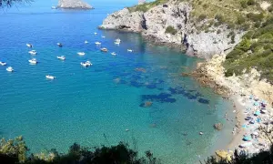 La cala del Gesso, Monte Argentario, offre un mare cristallino con barche all'ancora e una spiaggia affollata di bagnanti sotto ombrelloni colorati.