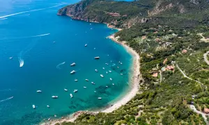 Vista aerea di una baia con spiagge dorate e acque cristalline, un vero gioiello della cartina della Toscana. Diverse imbarcazioni solcano il mare blu intenso.