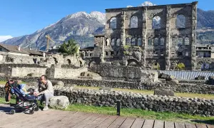 Un uomo con un passeggino ammira le rovine romane, uno dei **paesi più belli della Valle d'Aosta**, con montagne innevate sullo sfondo.