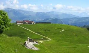 Un sentiero delle malghe serpeggia tra verdi colline, conducendo a un gruppo di rustici edifici con tetti rossi. In primo piano, ruderi di pietra e un piccolo stagno.