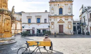 Un uomo seduto su una panchina in una piazza, con una bicicletta accanto. Architettura barocca e scorci dei borghi più belli del Salento.