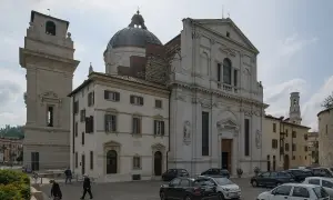 La piazza di San Giorgio a Verona, con la chiesa imponente, la torre campanaria e alcune persone che passeggiano tra le auto parcheggiate.