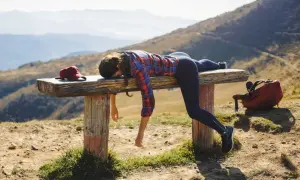 Donna esausta si riposa su una panca in montagna dopo un'intensa attività. Zaino e cappellino rossi accanto a lei.