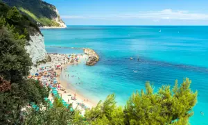 Spiaggia affollata con ombrelloni colorati e gente che prende il sole, bagnata da un **abruzzo mare bello** e cristallino, con scogliere bianche e vegetazione rigogliosa.