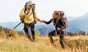 Coppia di escursionisti in montagna si aiutano a salire un pendio erboso, con zaini e bastoncini da trekking.