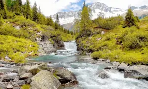 Cascata alpina e torrente cristallino, perfetti per gite in Trentino in giornata. Montagne innevate sullo sfondo.