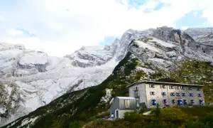 Un rifugio in pietra con persiane blu si staglia su un pendio verdeggiante, sotto imponenti montagne rocciose. Un esempio di rifugi in Veneto.