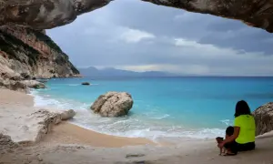 Vista dalla grotta sulla spiaggia di Cala Goloritzè, raggiungibile con un **percorso a piedi**. Una donna con un cane ammira il mare turchese.