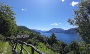 Il sentiero del viandante si snoda tra alberi verdi, con una vista mozzafiato su un lago azzurro e montagne sullo sfondo. Una piccola casa con tetto in pietra si intravede.
