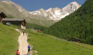 Escursionisti e ciclisti percorrono un sentiero nel **Val di Fosse**, tra baite in legno e montagne innevate.