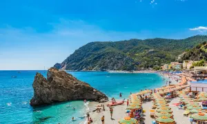 Spiaggia affollata sulla Riviera di Levante, con ombrelloni colorati, gente che nuota e montagne verdi sullo sfondo.