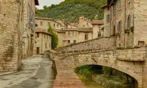 Antica via acciottolata con un ponte in pietra su un ruscello, circondata da case in pietra. Un'immagine perfetta dei borghi da visitare in Umbria.