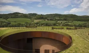 Un cerchio di metallo arrugginito emerge dal verde delle colline del Chianti, con vigneti che si estendono all'orizzonte sotto un cielo azzurro.