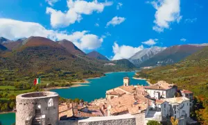 Borgo medievale con torre e lago turchese, un incantevole scorcio di cosa vedere in Abruzzo, tra montagne autunnali e cielo azzurro.