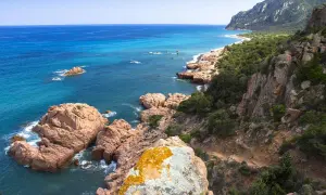 Scogliere rosa e mare turchese sulla costa ovest Sardegna. Un sentiero si snoda tra rocce e macchia mediterranea verso una spiaggia isolata.
