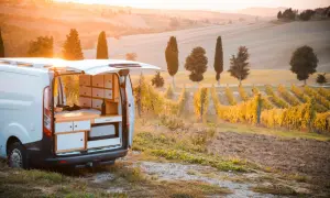 Un camper in un vigneto al tramonto, con dolci colline toscane sullo sfondo. L'interno del camper è visibile, con un letto e mobili.