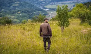 Uomo con fucile attraversa prati fioriti, godendosi la vista dei passi di montagna e della valle sottostante.