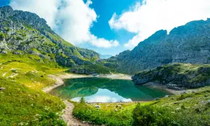 Sentiero in montagna tra verdi prati e rocce, con un lago alpino che riflette il cielo azzurro e nuvole bianche. Splendidi paesaggi naturali.