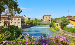 Un incantevole borgo da visitare in Veneto, con un fiume che scorre tra case antiche e una torre medievale. Fiori colorati incorniciano la scena.