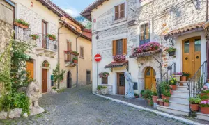 Cobblestone street in a medieval Italian village with stone buildings, balconies, and potted flowers.