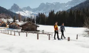 Coppia in cammino tra baite innevate e montagne maestose, un'esperienza di escursioni invernali in Trentino.