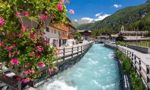 Fiori rosa adornano un vaso, affacciandosi su un torrente azzurro che attraversa i pittoreschi paesi del Val d'Ayas, con montagne innevate sullo sfondo.