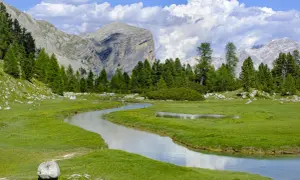 Un sereno sentiero di montagna serpeggia tra prati verdi e un ruscello cristallino, con imponenti vette sullo sfondo.