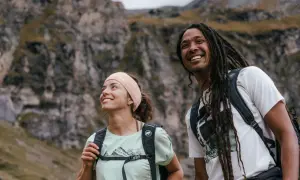 Coppia sorridente in montagna, con zaini e abbigliamento montagna estate perfetto per l'avventura.