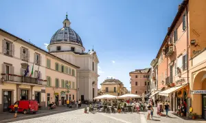 Piazza a Castel Gandolfo, dove si trova il Palazzo Apostolico. Edifici colorati, una chiesa con cupola e tavolini all'aperto animano la piazza.