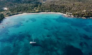 Vista aerea di una baia con acque cristalline, una barca a vela e una spiaggia dorata. Un assaggio delle migliori spiagge Costa Smeralda.