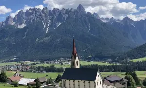 Chiesa con campanile rosso in primo piano, sullo sfondo le maestose vette delle Dolomiti e i **paesi più belli** della **Val Pusteria**.