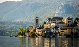 A picturesque Italian village with colorful buildings and a tall bell tower sits on the edge of a calm lake, with tree-covered mountains in the background.