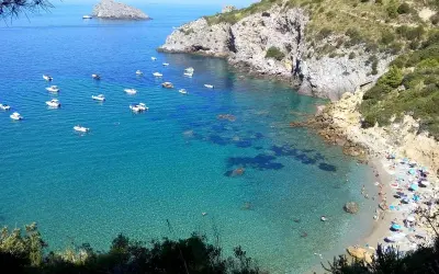 La cala del Gesso, Monte Argentario, offre un mare cristallino con barche all'ancora e una spiaggia affollata di bagnanti sotto ombrelloni colorati.