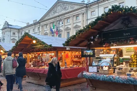 Mercatini di Natale Aosta: bancarelle addobbate con luci e ghirlande, prodotti tipici e gente che passeggia. Atmosfera magica.