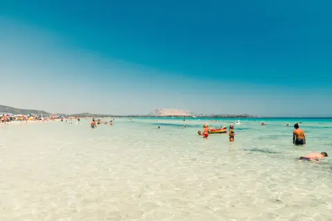 Spiaggia affollata in Sardegna, tra le zone più belle, con gente che nuota e si rilassa nell'acqua cristallina.