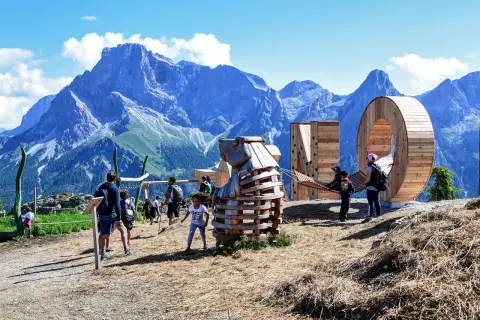 Un'estate a San Martino di Castrozza: bambini giocano in un parco avventura con sculture in legno e montagne maestose sullo sfondo.