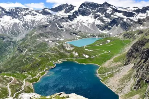 Laghi alpini e strada tortuosa sul colle del Nivolet, con montagne innevate sullo sfondo.
